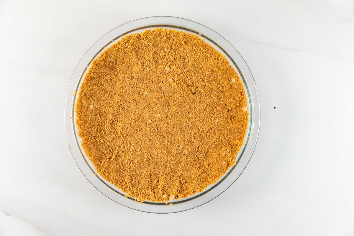 A pie dish filled with a smooth, evenly pressed graham cracker crust resting on a white countertop.