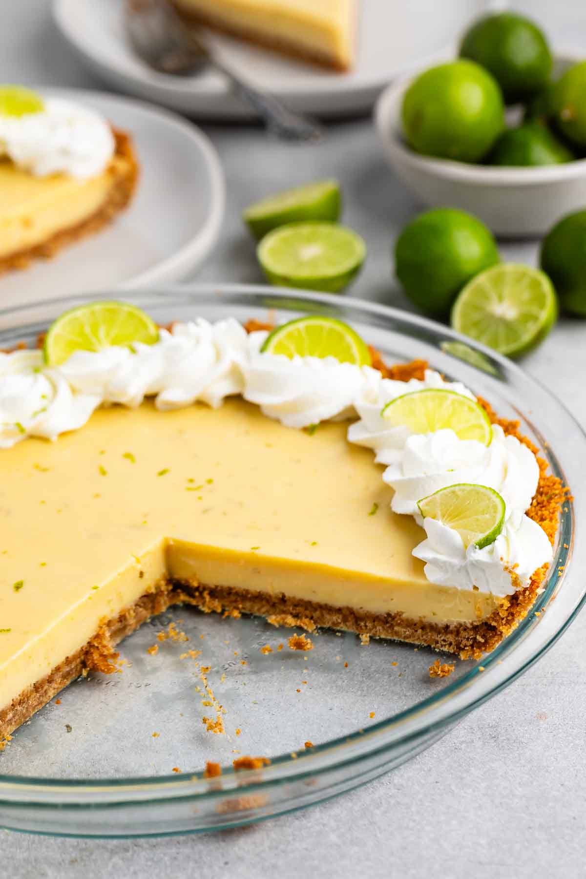 A key lime pie with a slice removed, showing its creamy filling and crumbly crust. The pie is garnished with whipped cream and lime slices. Whole limes and a plate with a slice are in the background.