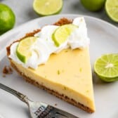 A slice of key lime pie on a white plate, topped with whipped cream and lime slices. A fork rests beside it. Halved and whole limes are in the background.