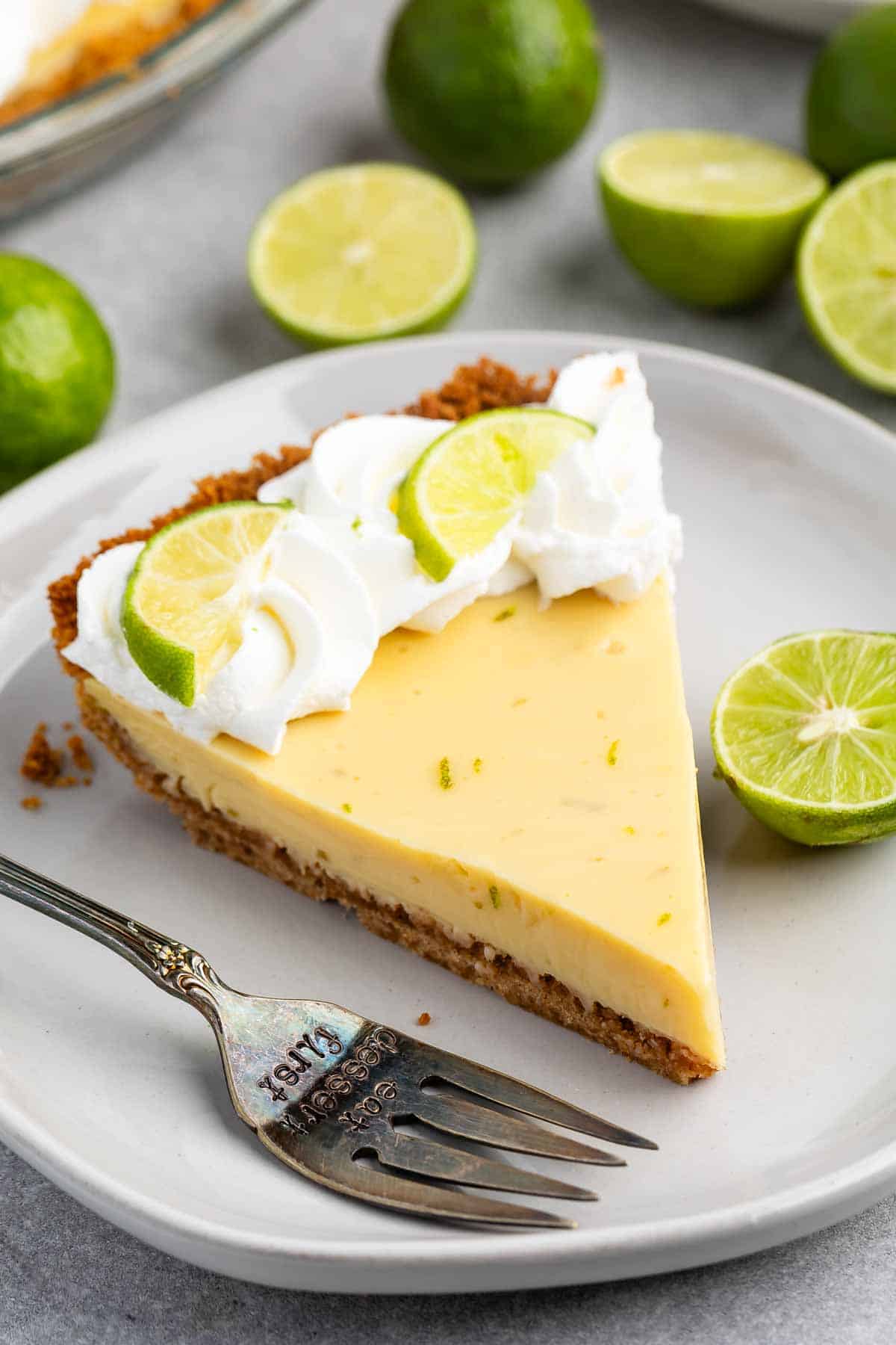 A slice of key lime pie on a white plate, topped with whipped cream and lime slices. A fork rests beside it. Halved and whole limes are in the background.