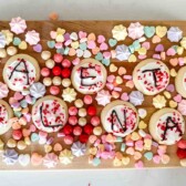 A wooden board displaying sugar cookies with letters spelling VACAY VIBES, surrounded by colorful candies, heart-shaped candies, and pastel-colored meringues.