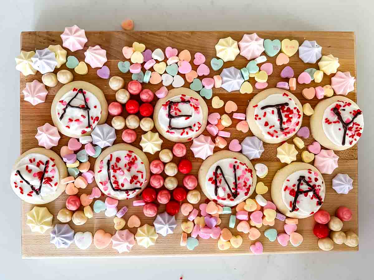 A wooden board displaying sugar cookies with letters spelling VACAY VIBES, surrounded by colorful candies, heart-shaped candies, and pastel-colored meringues.