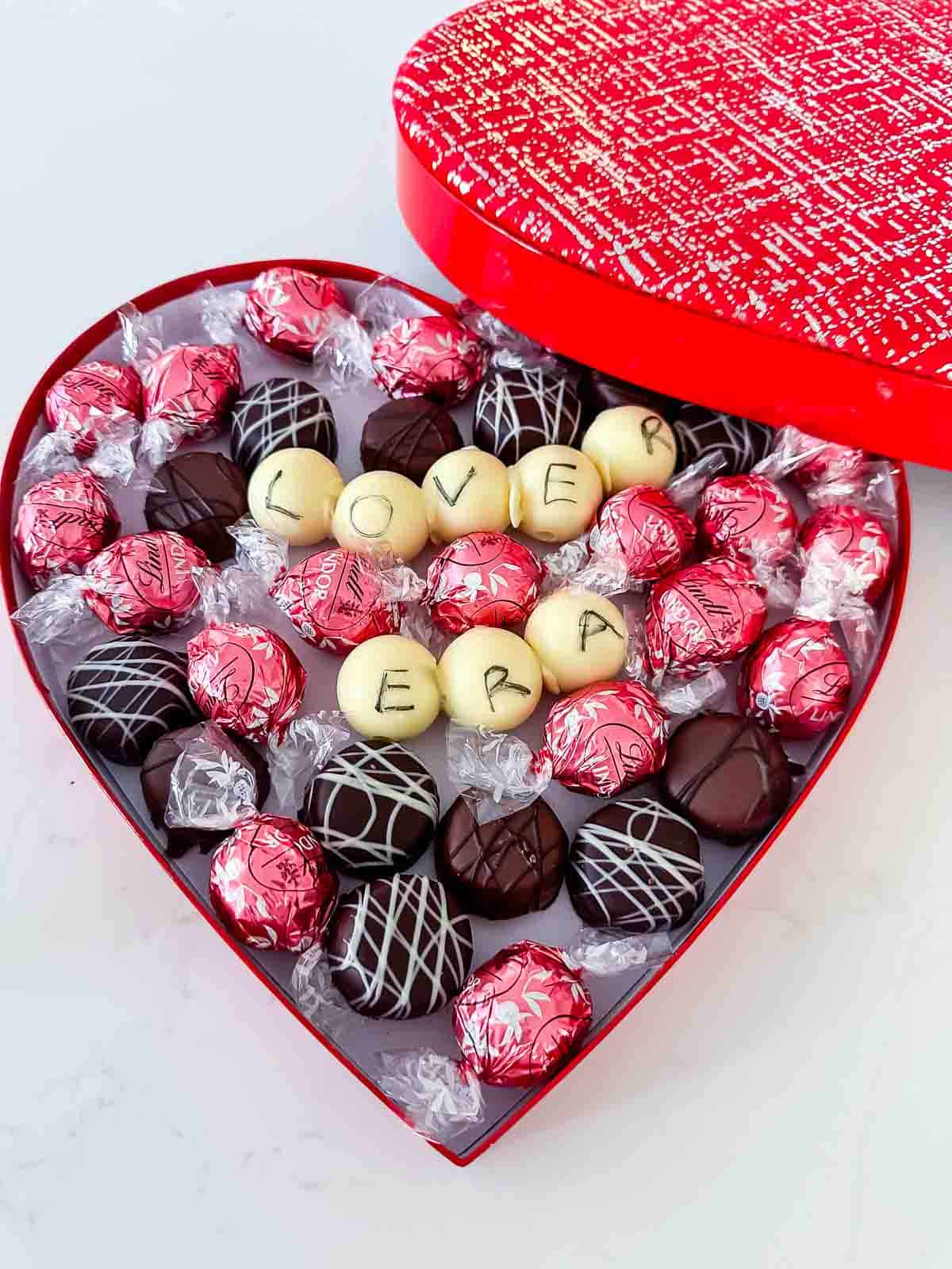 A heart-shaped red box filled with assorted chocolates, including foil-wrapped ones and truffles with decorative drizzles. In the center, white chocolates spell out LOVE and ERA.