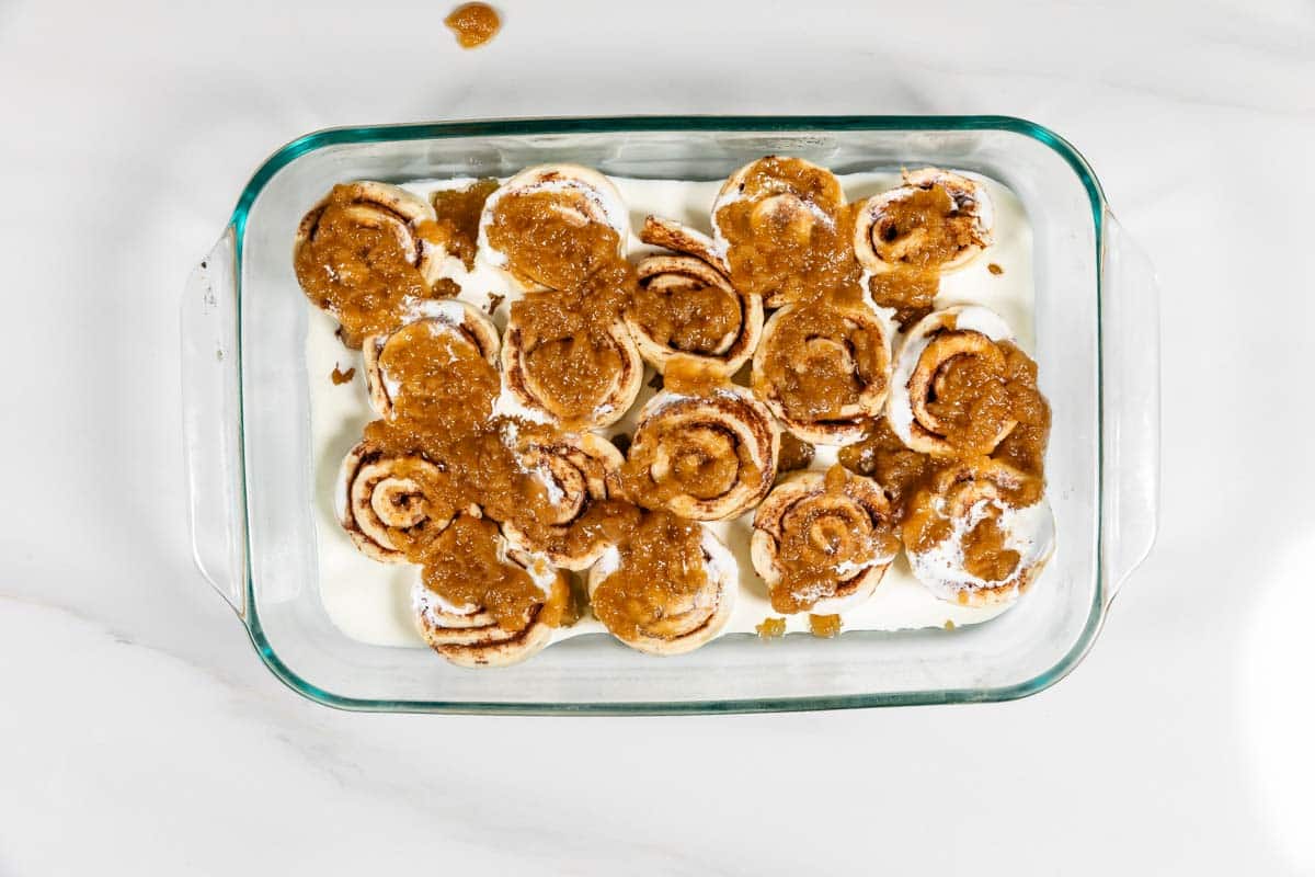 A glass baking dish filled with unbaked cinnamon rolls topped with brown sugar and butter mixture, resting on a white countertop. The rolls are arranged closely together, ready for baking.