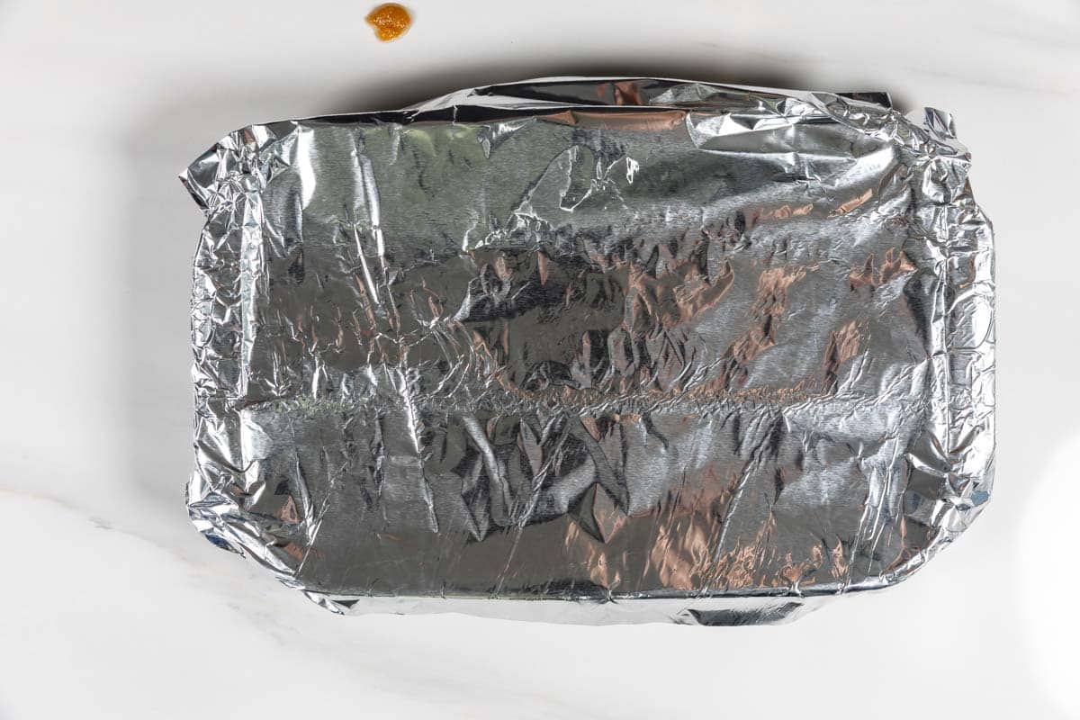 A rectangular dish covered with aluminum foil sits on a white surface. A small amount of sauce or residue can be seen near the top edge of the image.