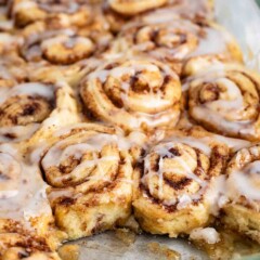 A glass baking dish filled with freshly baked cinnamon rolls, topped with a glossy glaze. Some rolls have a golden-brown crust and a spiral shape, with cinnamon filling visible. One roll is partially removed, revealing its soft, fluffy texture.