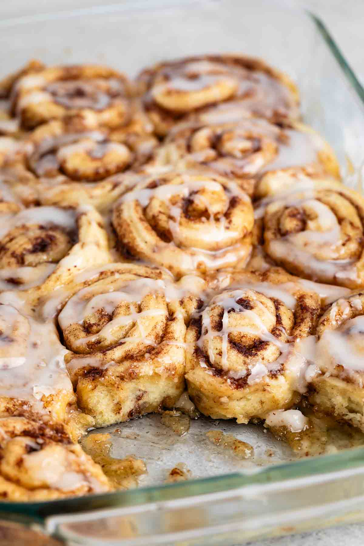 A glass baking dish filled with freshly baked cinnamon rolls, topped with a glossy glaze. Some rolls have a golden-brown crust and a spiral shape, with cinnamon filling visible. One roll is partially removed, revealing its soft, fluffy texture.