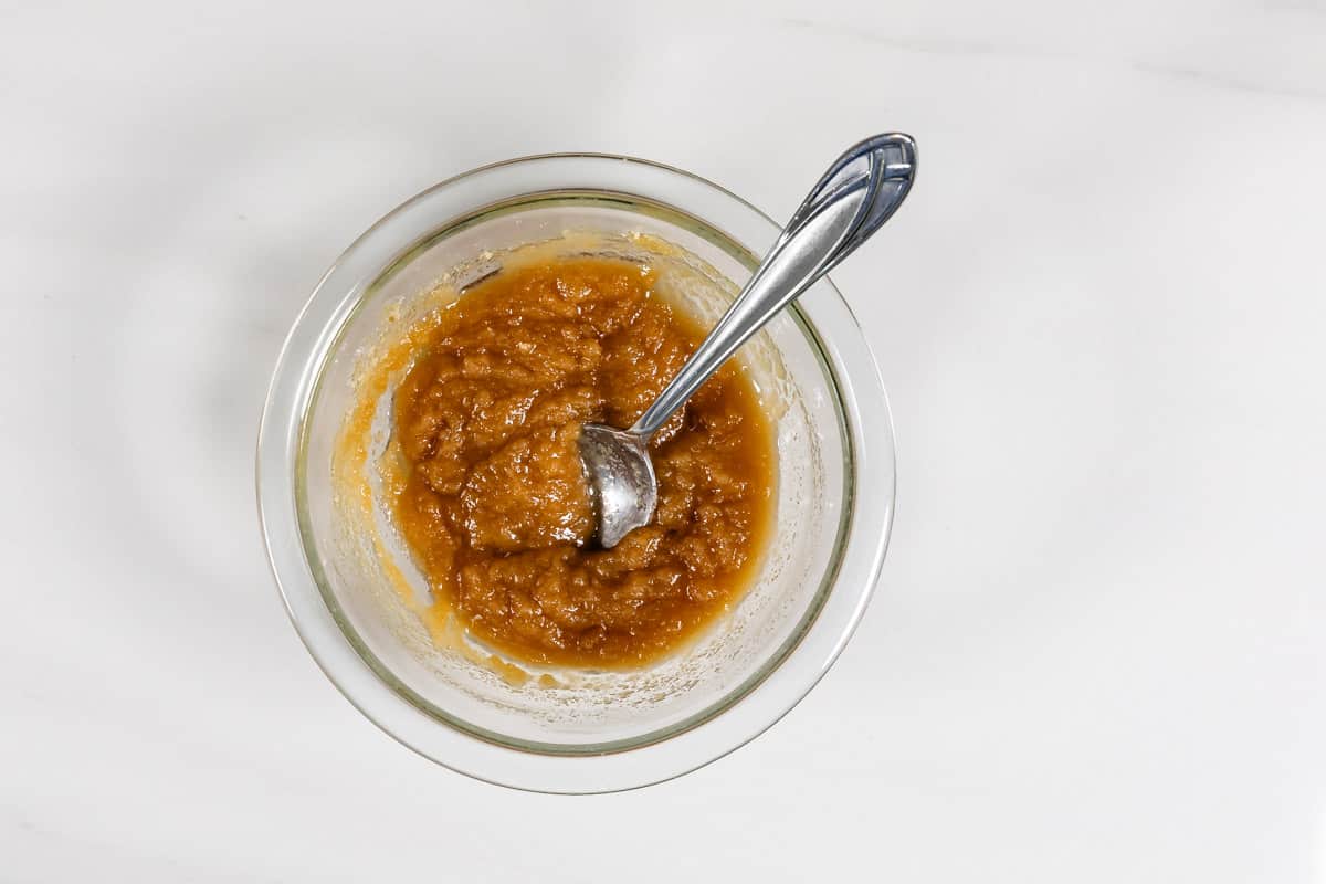 A glass bowl filled with applesauce, with a metal spoon resting inside, on a white surface.