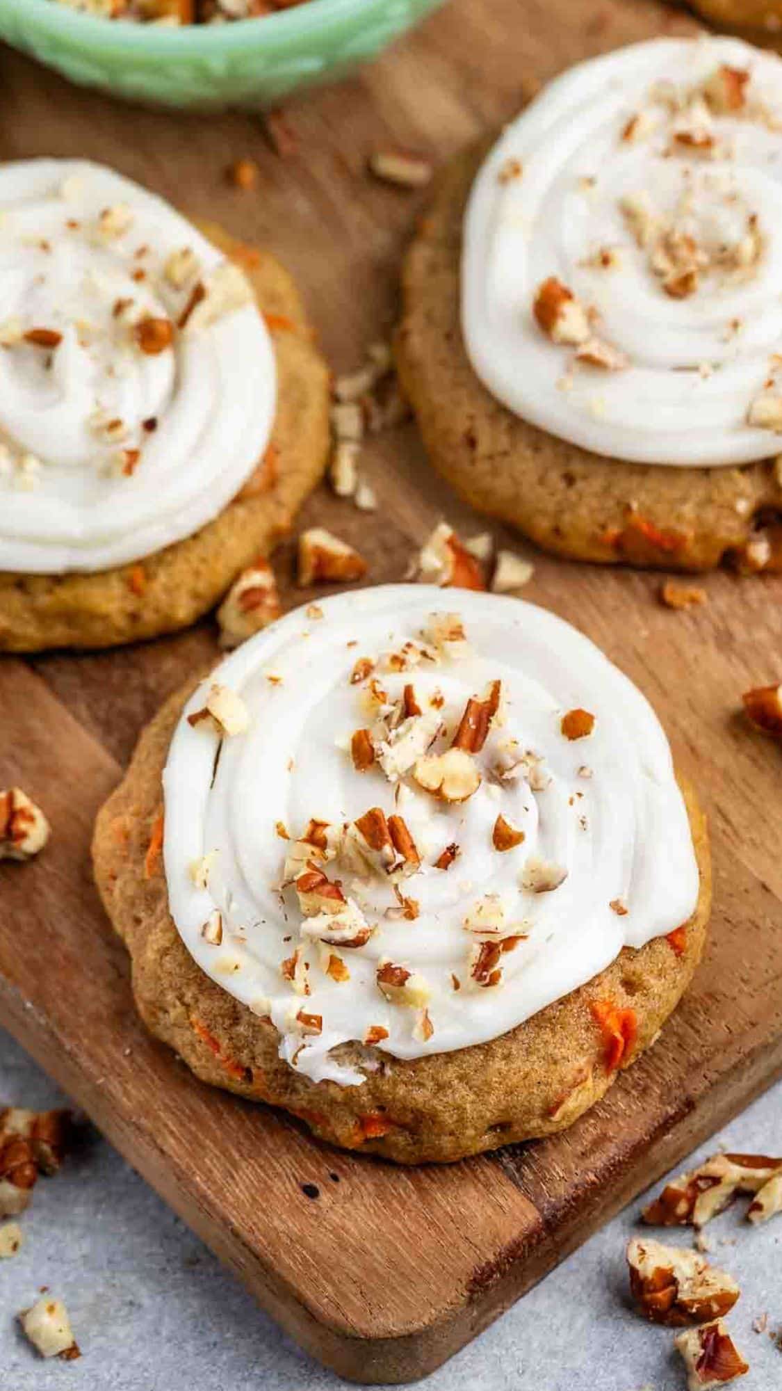 Three carrot cake cookies with white frosting and chopped nuts are arranged on a wooden board. Some nuts are sprinkled around the cookies. A green bowl is partially visible in the background.