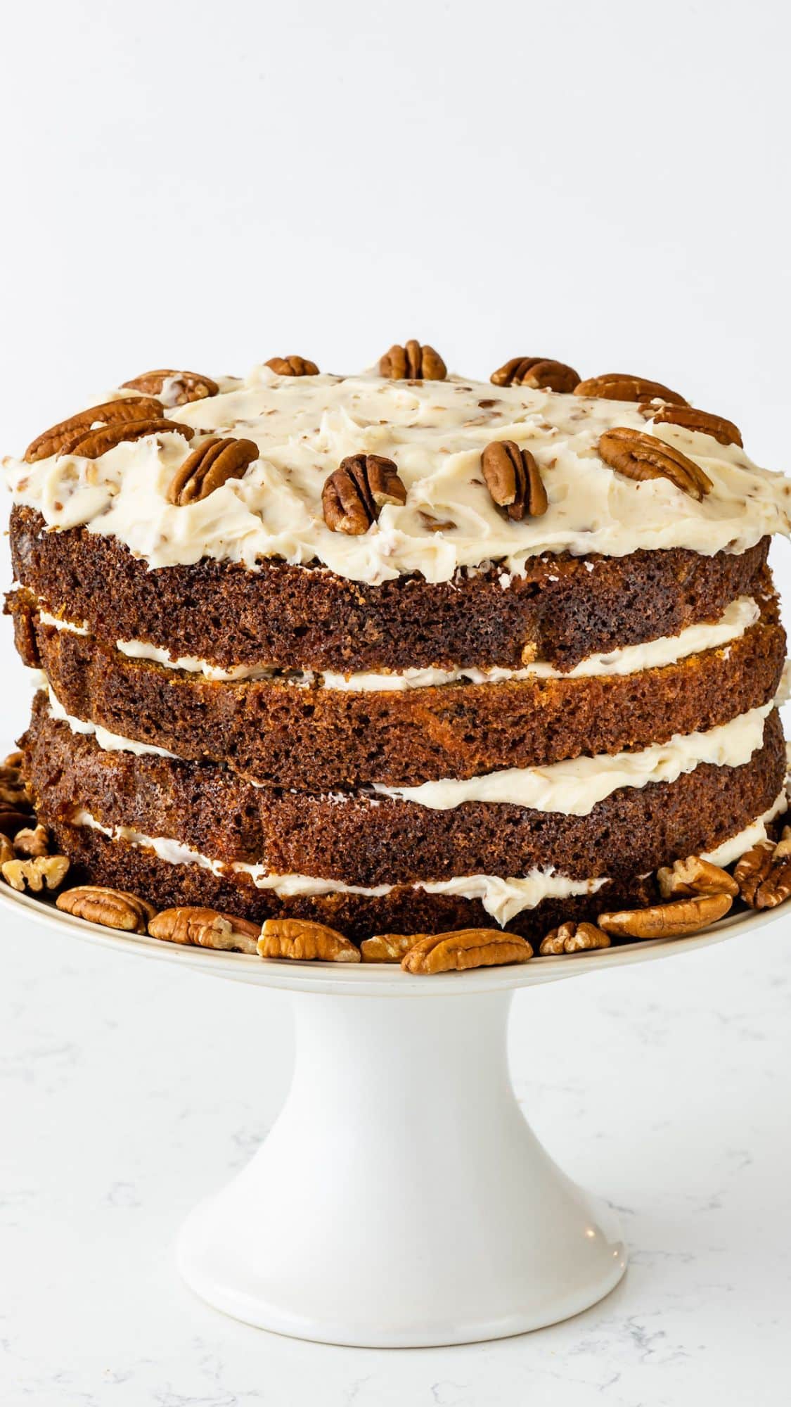 A layered carrot cake with cream cheese frosting, topped and surrounded by pecan halves, displayed on a white cake stand against a light background.