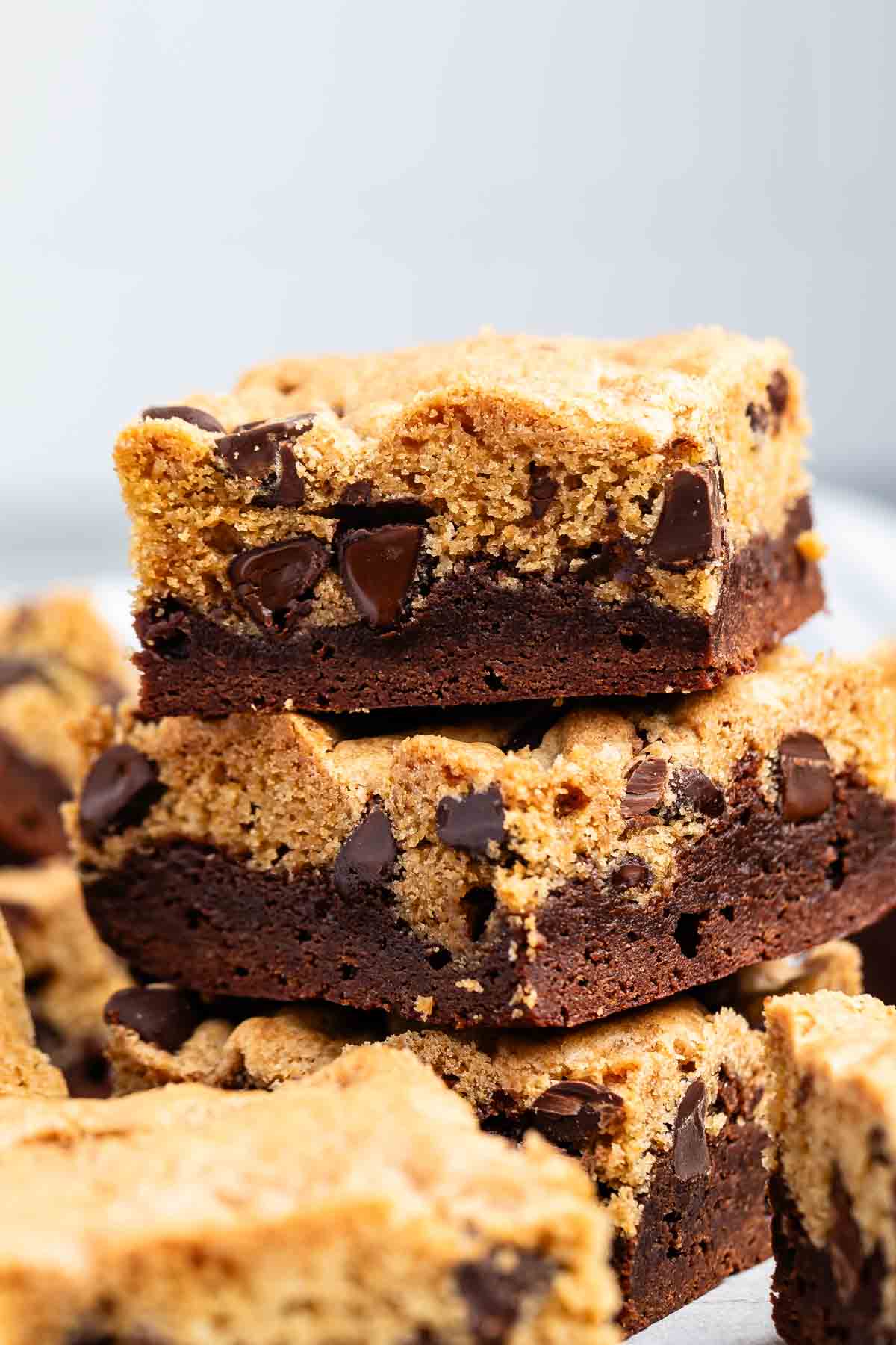 Stacked squares of brookies, a dessert combining chocolate chip cookies and brownies, are shown. The top layer is cookie dough with chocolate chips, while the bottom layer is brownie.