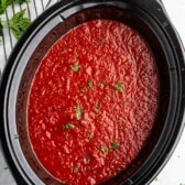 A slow cooker filled with vibrant red tomato sauce, garnished with chopped green herbs. A striped kitchen towel and fresh parsley are partially visible on the side.