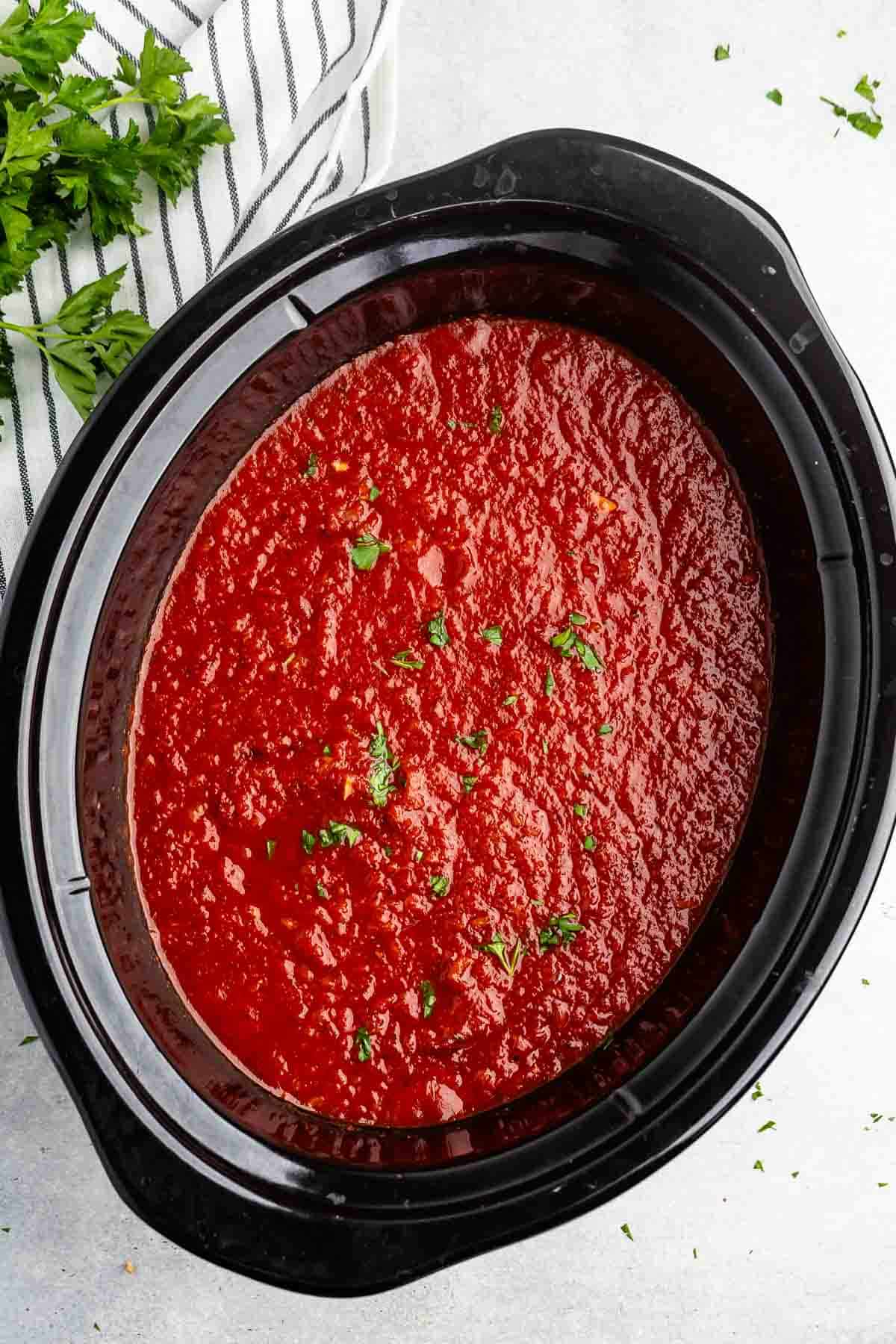 A slow cooker filled with vibrant red tomato sauce, garnished with chopped green herbs. A striped kitchen towel and fresh parsley are partially visible on the side.