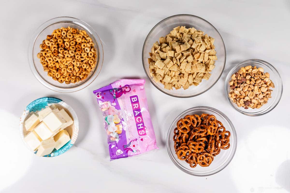 Various snack ingredients are displayed in bowls on a white surface, including cheerios, rice cereal squares, mixed nuts, pretzels, and white candy melts. A pink package labeled Brachs is in the center.