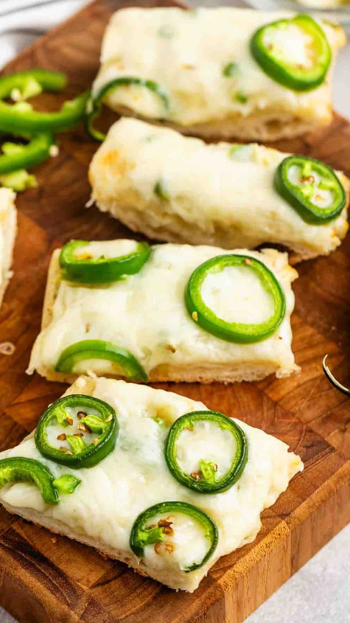 Slices of bread topped with melted cheese and jalapeño slices are arranged on a wooden cutting board. Additional jalapeño slices are scattered nearby.