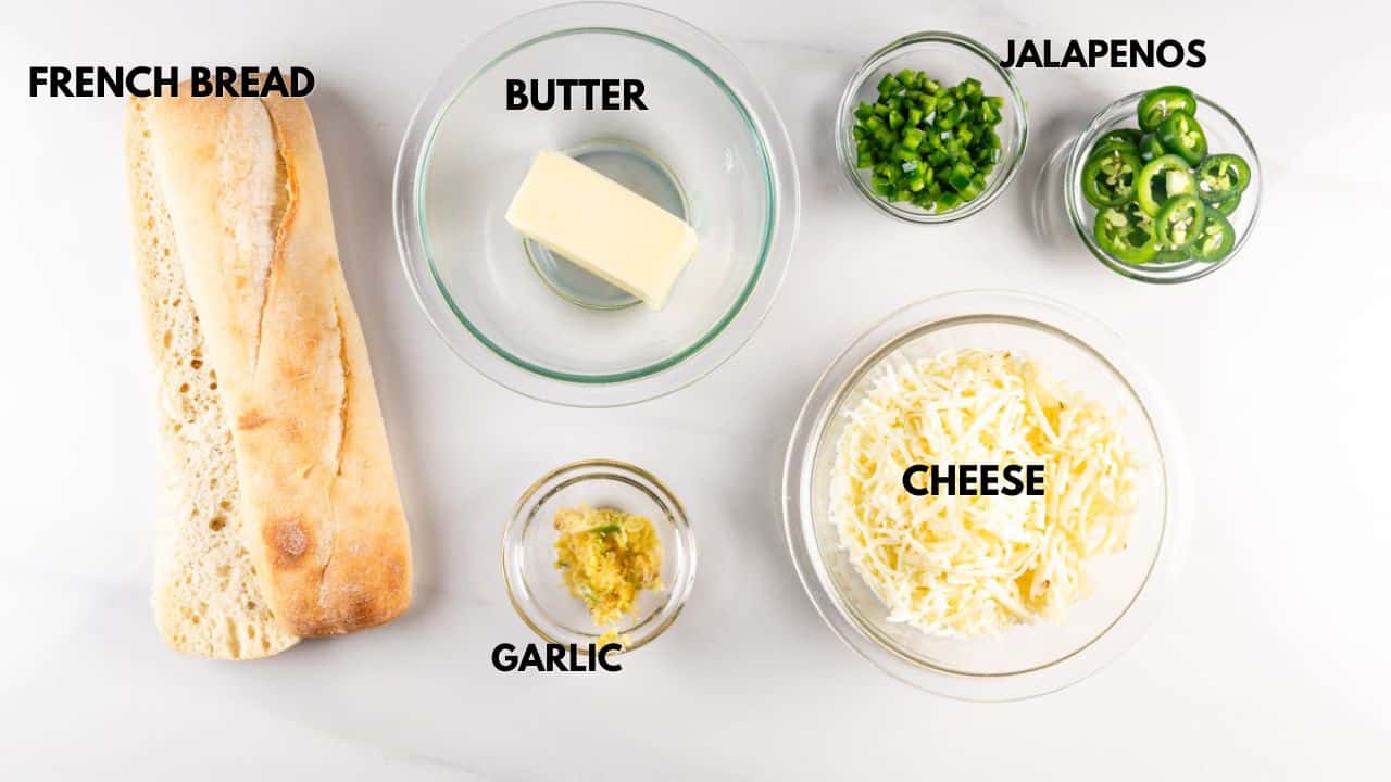 Ingredients on a white surface: a loaf of French bread, a bowl of butter, chopped and sliced jalapeños, a bowl of cheese, and a small bowl of minced garlic. Labels identify each item.