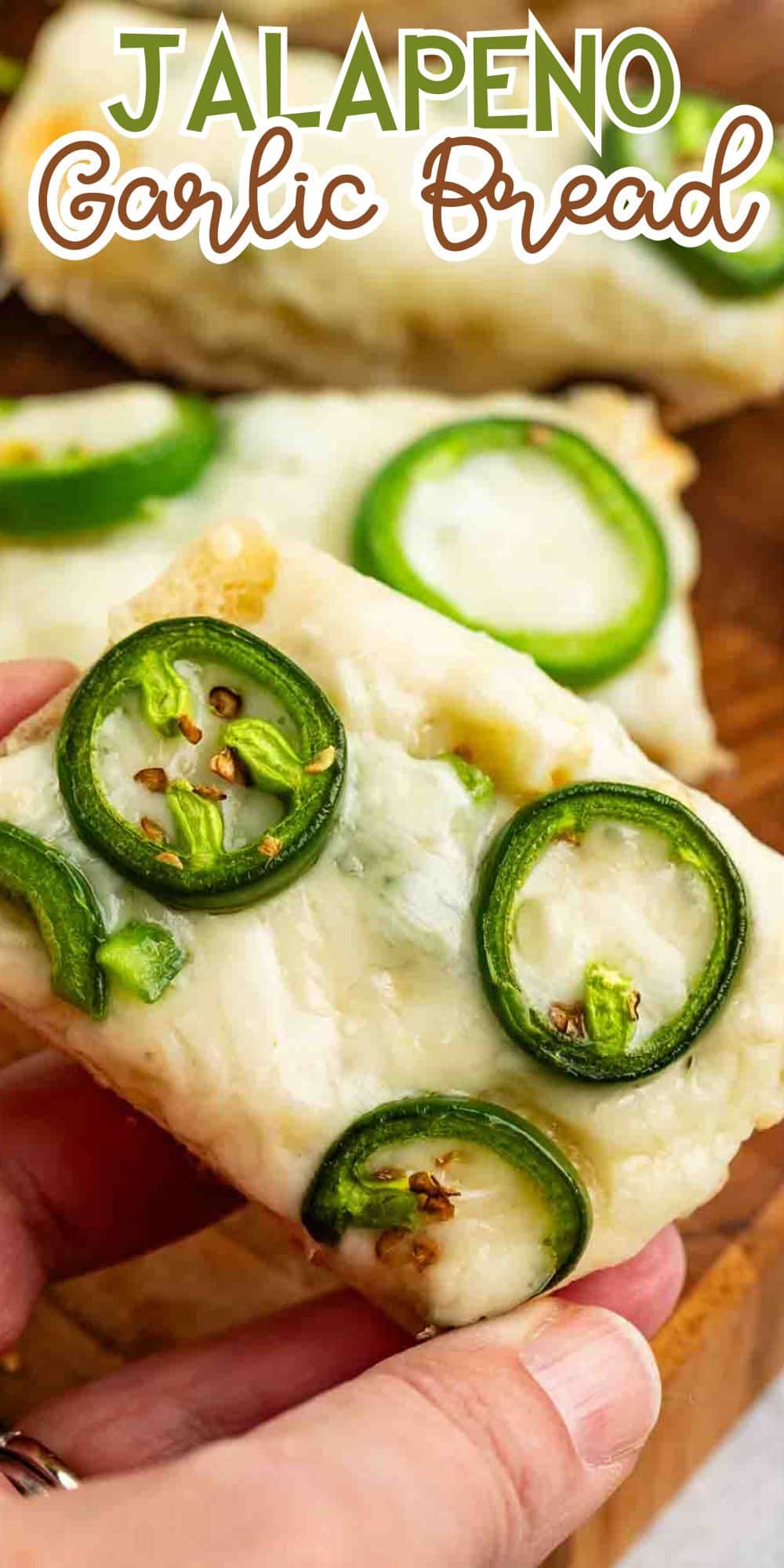 A close-up of a hand holding a slice of jalapeno garlic bread. The bread is topped with melted cheese and sliced jalapenos, with a creamy texture. More slices are visible on a wooden surface in the background. Text reads Jalapeno Garlic Bread.