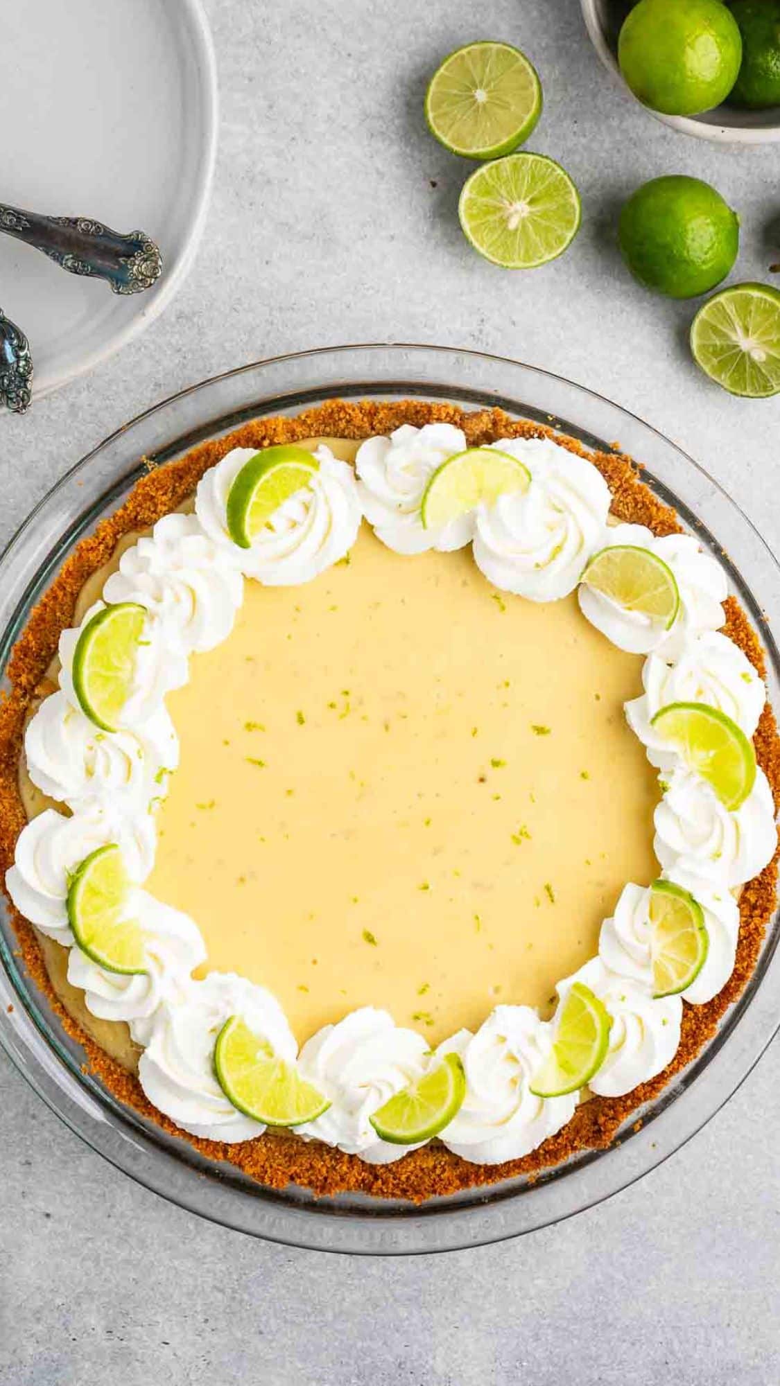 Key lime pie in a clear dish, topped with whipped cream and lime slices. A few whole limes and a gray plate with forks are in the background on a gray surface.