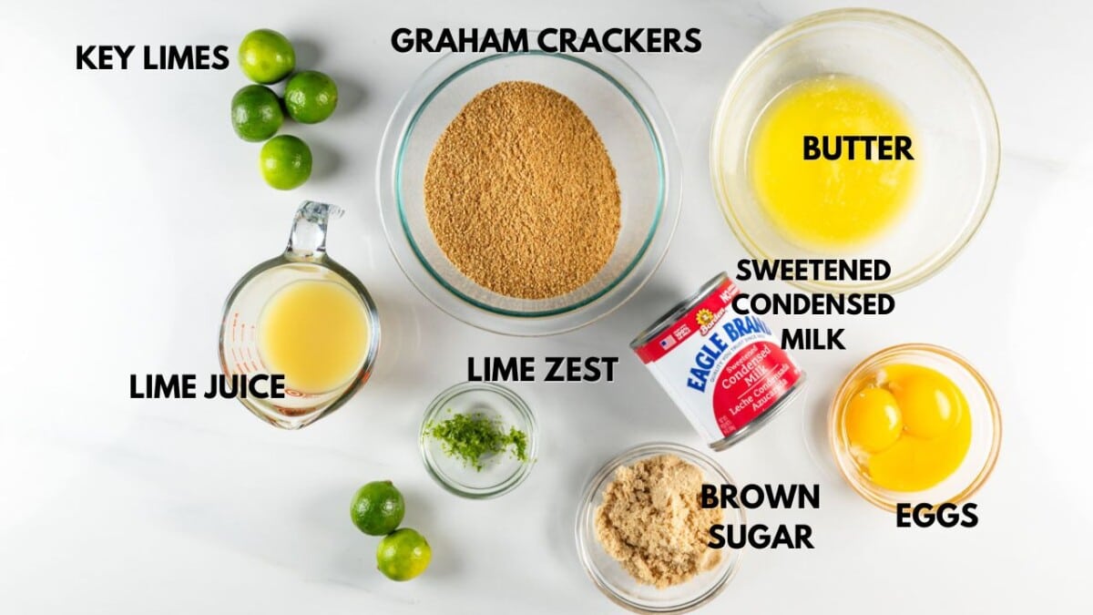 Ingredients for a recipe arranged on a white surface: a bowl of graham cracker crumbs, melted butter, sweetened condensed milk, eggs, brown sugar, lime juice, lime zest, and key limes. Each item is labeled.