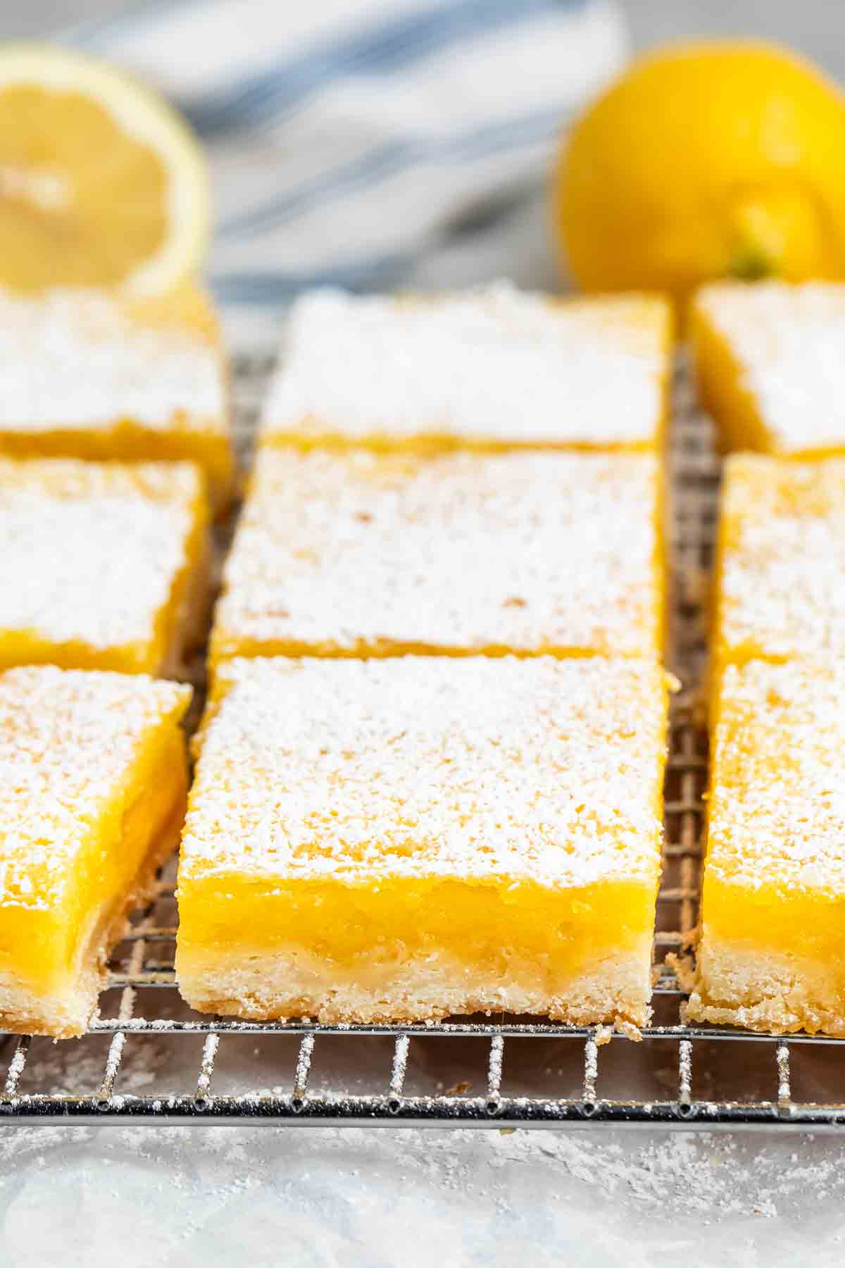 Close-up of lemon bars on a cooling rack, topped with powdered sugar. A fresh lemon is blurred in the background.