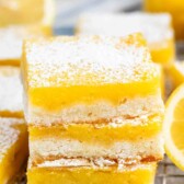 Close-up of lemon bars on a cooling rack, topped with powdered sugar. A fresh lemon is blurred in the background.