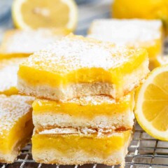 Close-up of lemon bars on a cooling rack, topped with powdered sugar. A fresh lemon is blurred in the background.