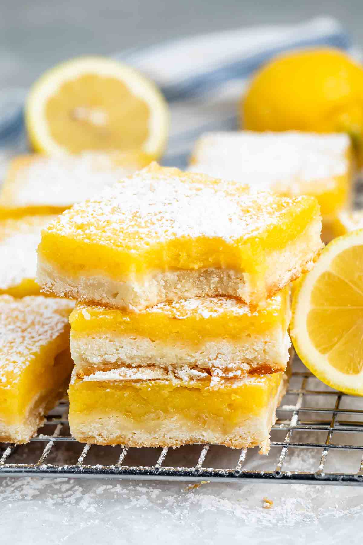 Close-up of lemon bars on a cooling rack, topped with powdered sugar. A fresh lemon is blurred in the background.