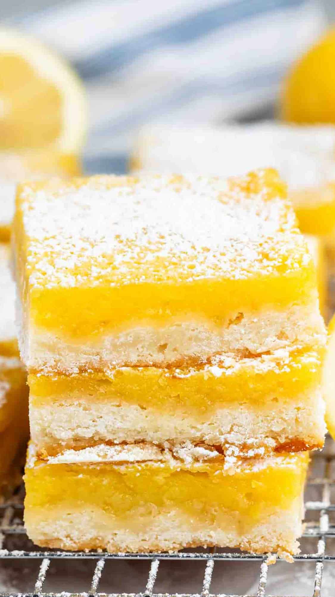 Three stacked lemon bars on a cooling rack, topped with powdered sugar. More lemon bars and a lemon are blurred in the background.