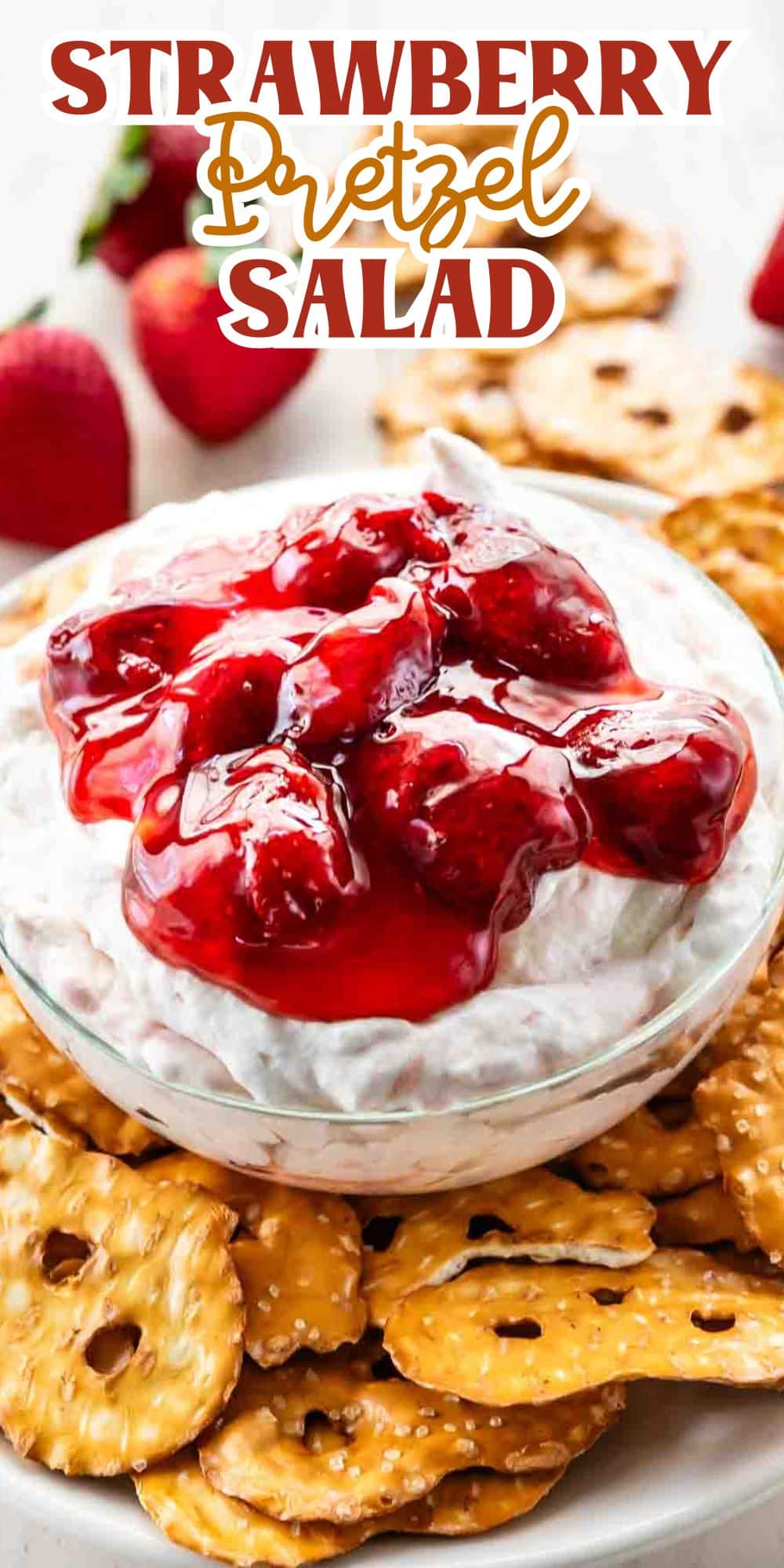 A glass bowl filled with strawberry pretzel salad is surrounded by pretzel crisps. The salad has a creamy base topped with a layer of strawberries in gelatin. Above the bowl, Strawberry Pretzel Salad is written in decorative text.