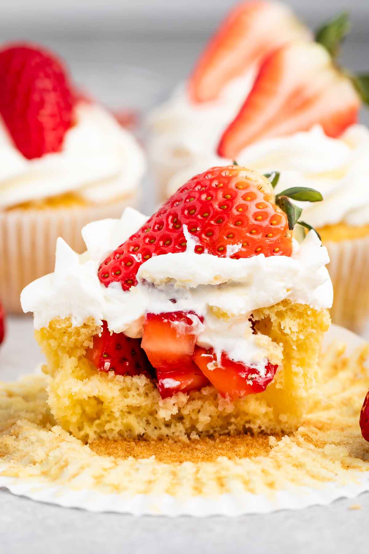 Vanilla cupcake with white frosting topped with a fresh strawberry, surrounded by more cupcakes.