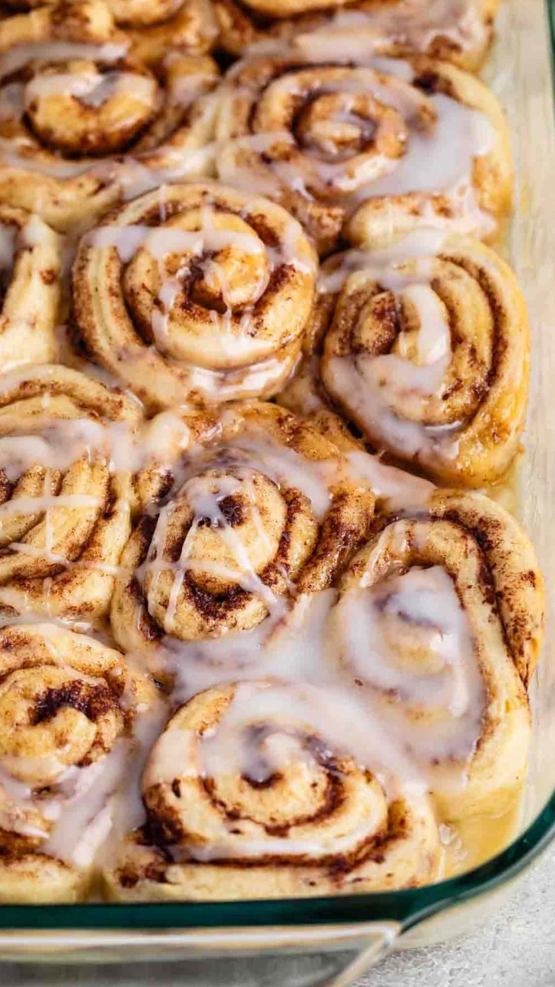 A close-up of freshly baked cinnamon rolls in a glass baking dish. The rolls are swirled with cinnamon and topped with a glossy layer of melting icing, giving them a warm and inviting appearance.