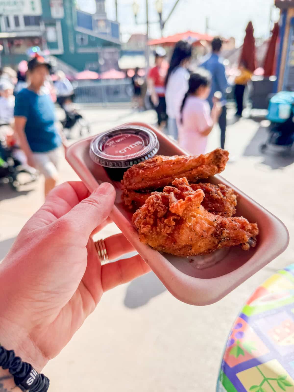 A hand holding a small rectangular tray with two pieces of fried chicken and a sauce cup. The background shows a bustling outdoor setting with people and colorful tables.