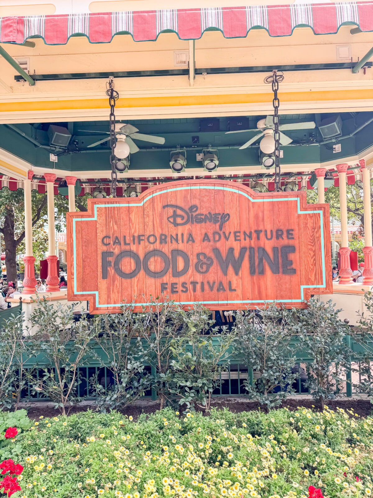 A wooden sign with Disney California Adventure Food & Wine Festival is displayed on a decorative structure with red and white striped canopy. Surrounding the structure are green plants and colorful flowers.