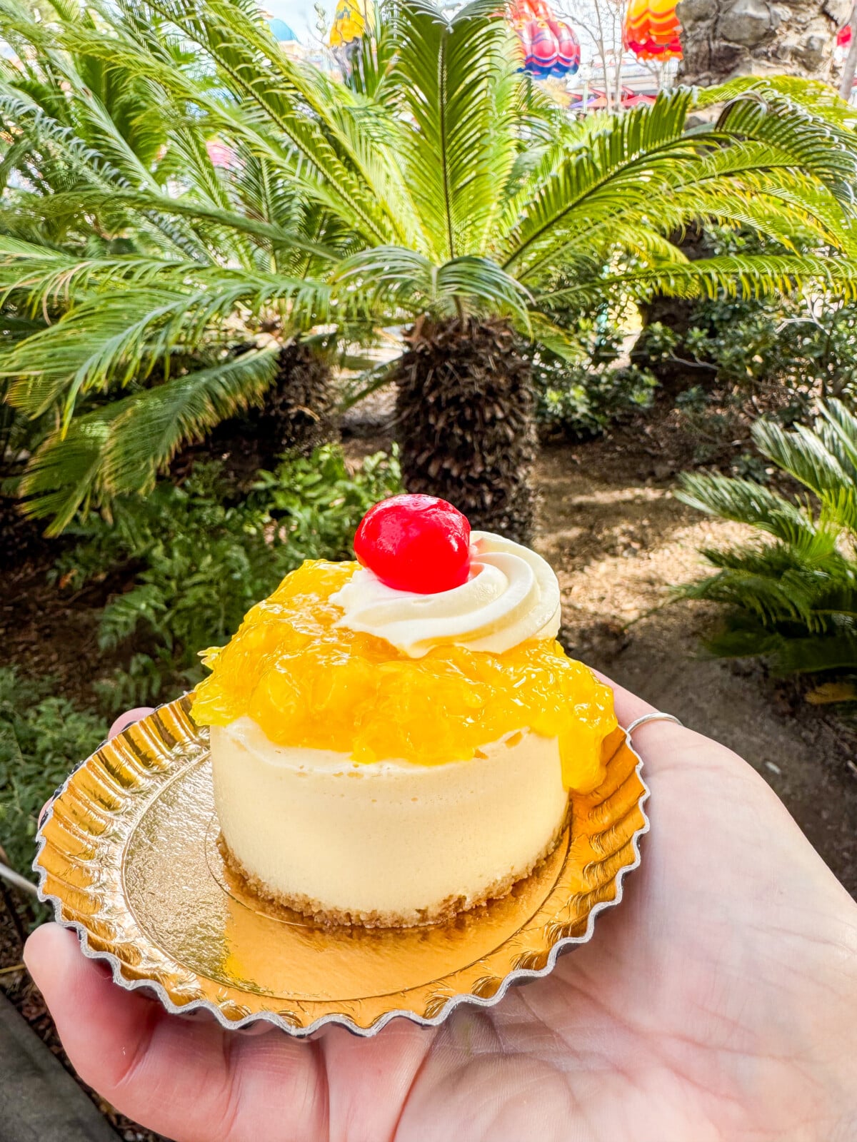 A hand holds a small cheesecake topped with vibrant yellow pineapple chunks and a red cherry, served on a gold plate. A background of lush green palm trees enhances the tropical vibe.