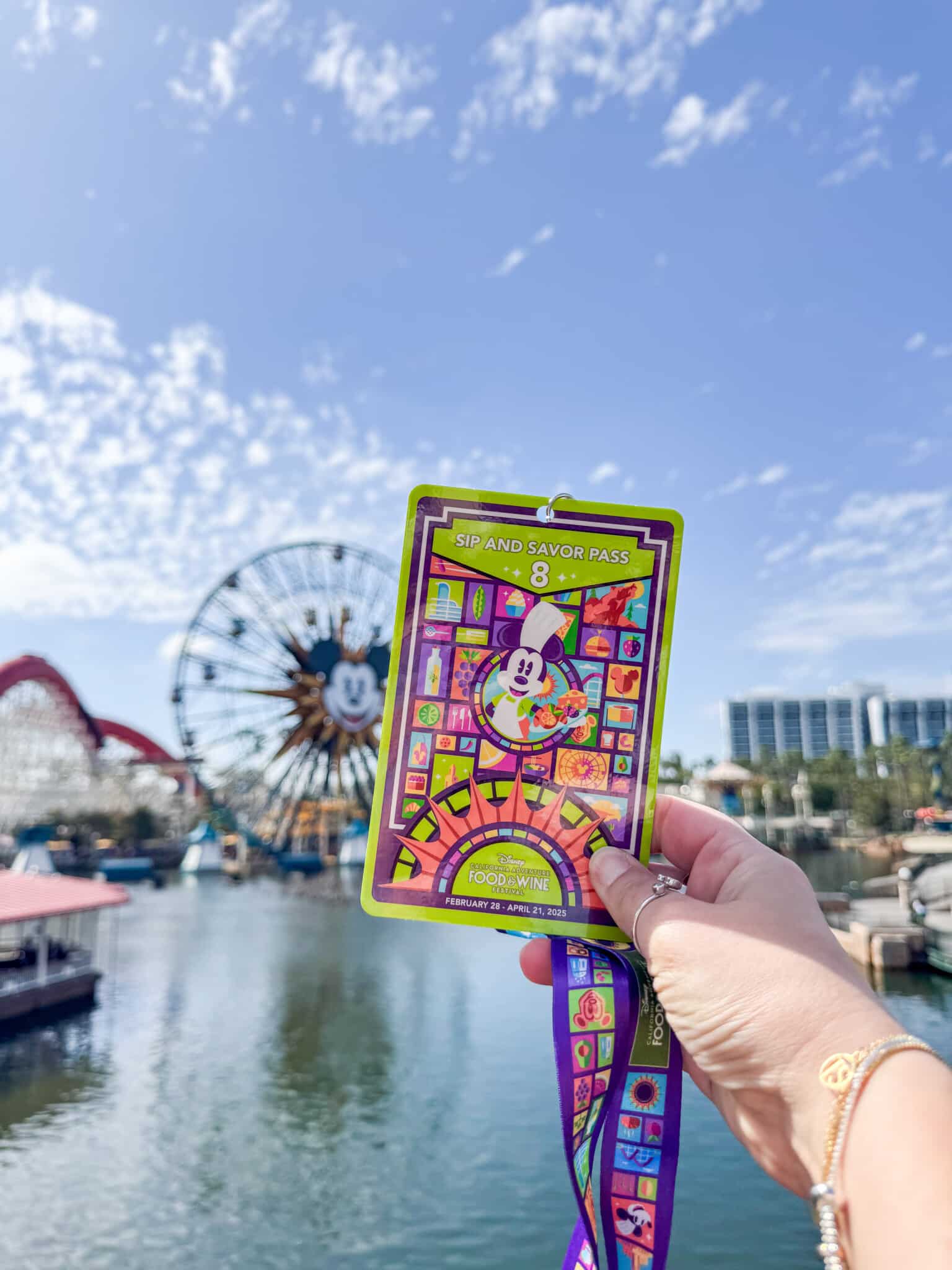 A hand holds up a colorful Sip and Savor Pass with an illustration of a cartoon character. In the background, theres an amusement park with a Ferris wheel featuring a similar cartoon characters face and a clear blue sky.