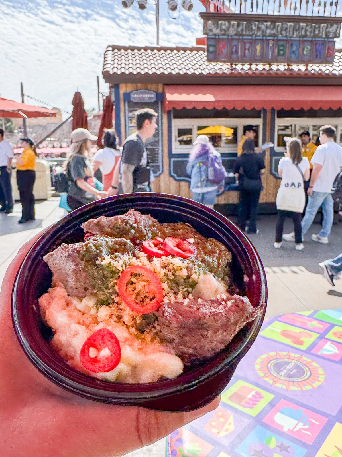 A bowl of seasoned meat placed over mashed potatoes, topped with chili slices and herbs. The dish is held up in front of a bustling outdoor food stall, with people standing and walking around on a sunny day.