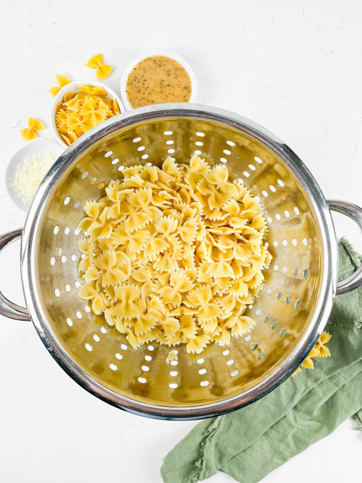 Cooked bowtie pasta in a metal colander on a white surface. Nearby are small bowls with dressing and grated cheese. A few uncooked bowtie pasta pieces and a green cloth are also visible.