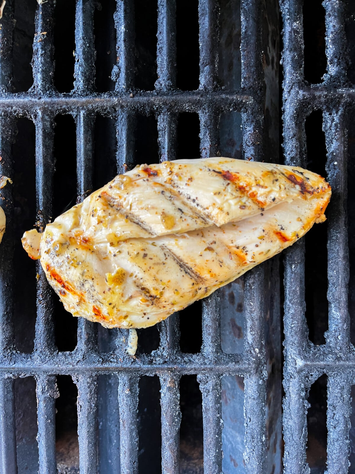 A seasoned chicken breast is grilling on a metal barbecue grate. The meat has visible grill marks and a slightly charred exterior, indicating it is being cooked over an open flame. The grate is dark with some rust spots.