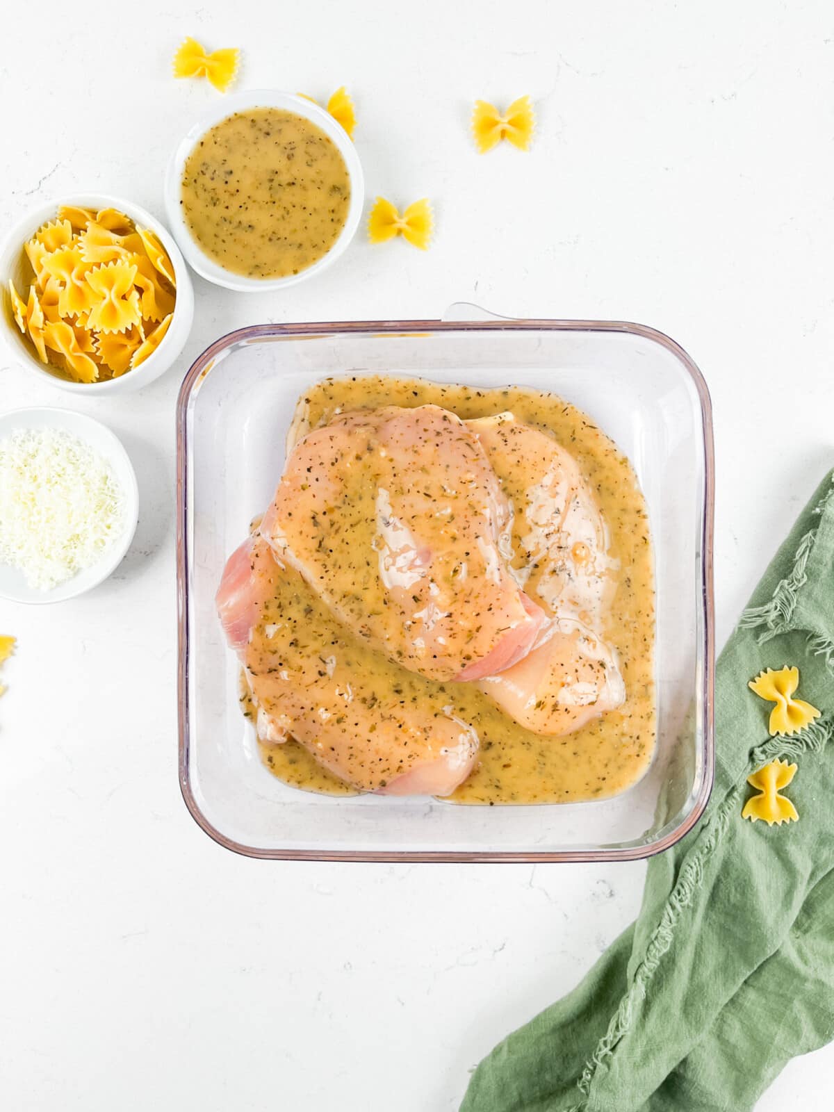 Raw chicken breast marinating in a glass dish with a creamy herb sauce. Surrounding the dish are small bowls containing uncooked bow tie pasta and grated Parmesan cheese. A green cloth and a few scattered pasta pieces are nearby on a white surface.