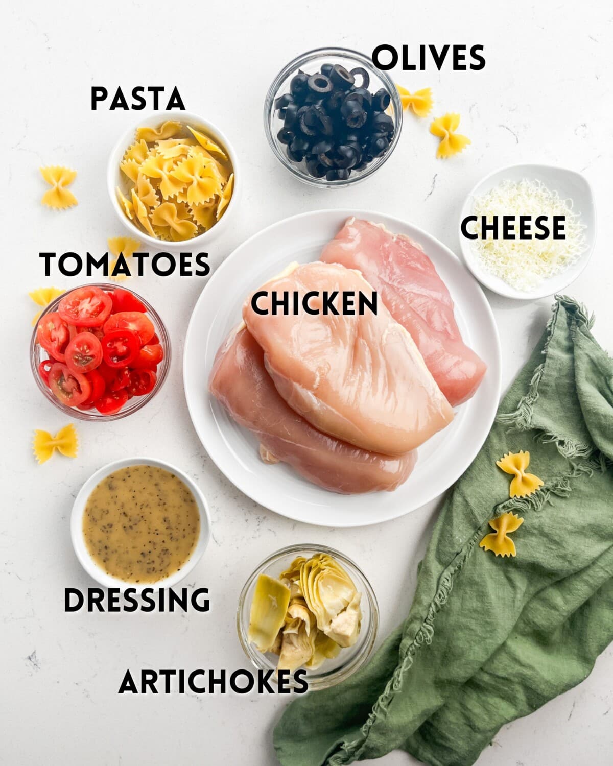 A collection of ingredients labeled for a dish: chicken breasts on a plate, and bowls of sliced olives, shredded cheese, cherry tomatoes, pasta, artichokes, and dressing, arranged on a white surface with a green cloth.