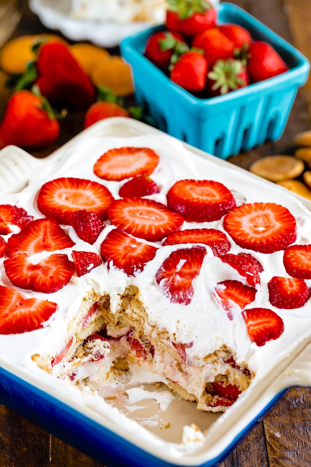 A dish with a partially eaten dessert topped with whipped cream and sliced strawberries. A basket of fresh strawberries is in the background alongside scattered cookies on a wooden surface.