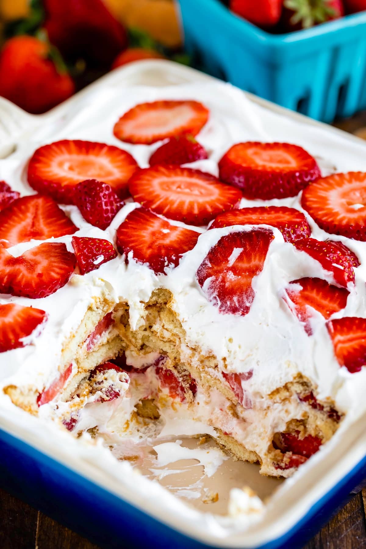 A close-up of a strawberry icebox cake in a rectangular dish. The cake has layers of whipped cream and sliced strawberries on top. A portion of the cake is missing, revealing its layered interior. Fresh strawberries are in the background.