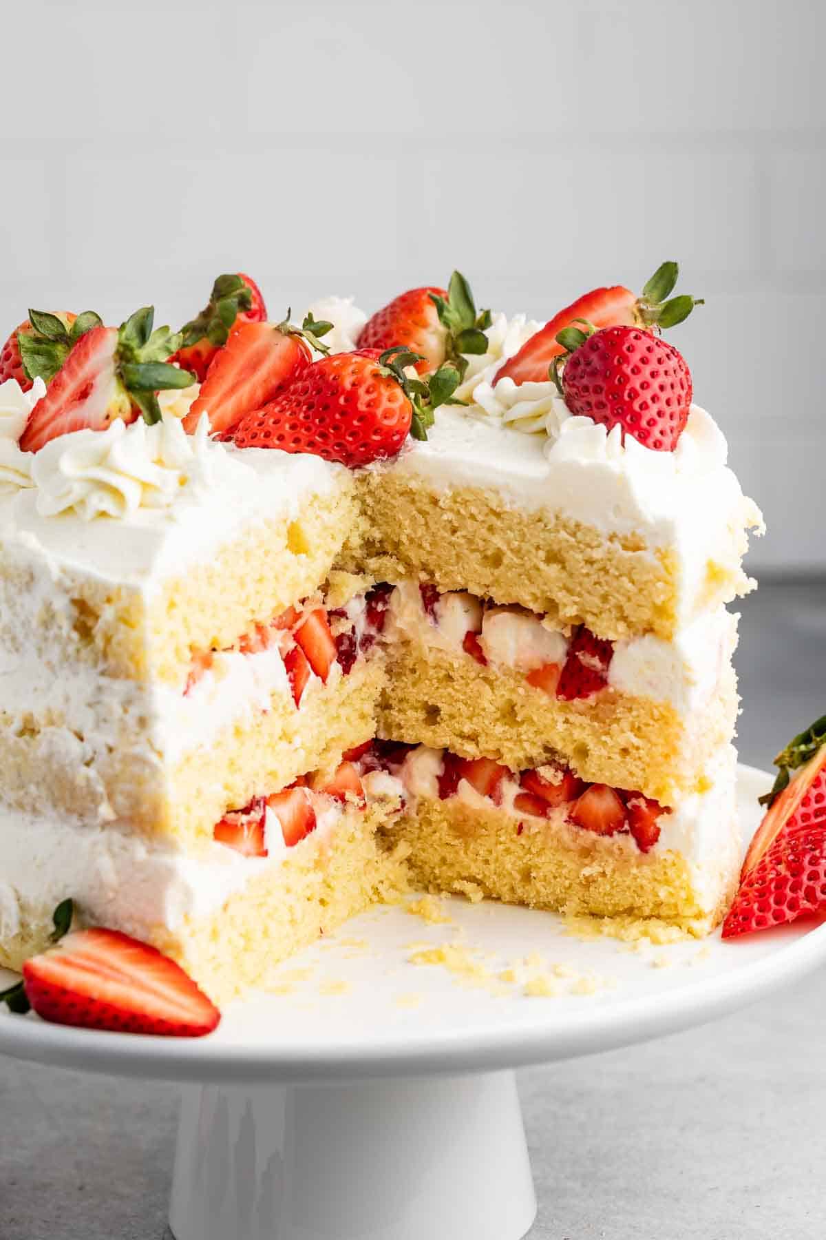 A sliced three-layer vanilla cake is topped with fresh strawberries and white frosting. Each layer contains cream and strawberry pieces. The cake is displayed on a white cake stand against a light gray background.