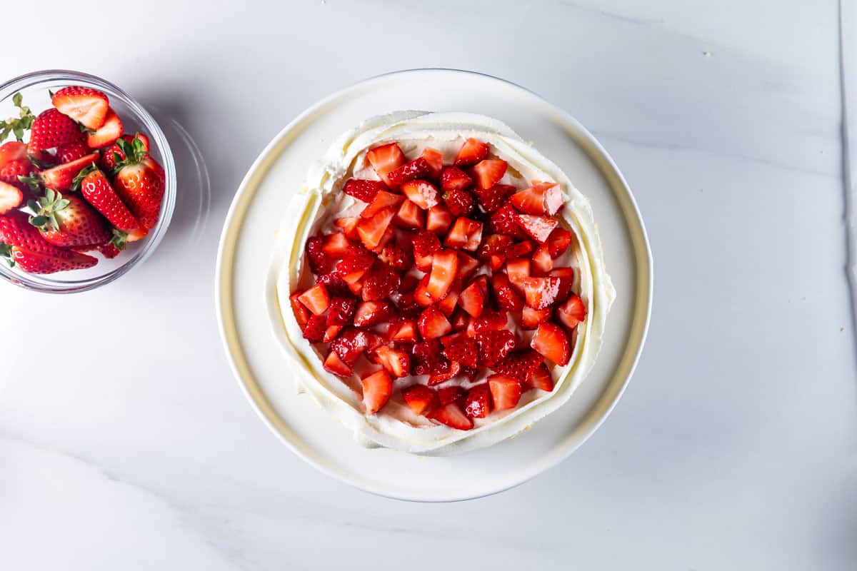 A round cake on a white plate topped with whipped cream and chopped fresh strawberries. To the left, a glass bowl filled with whole strawberries is visible. The background is a smooth, white surface.