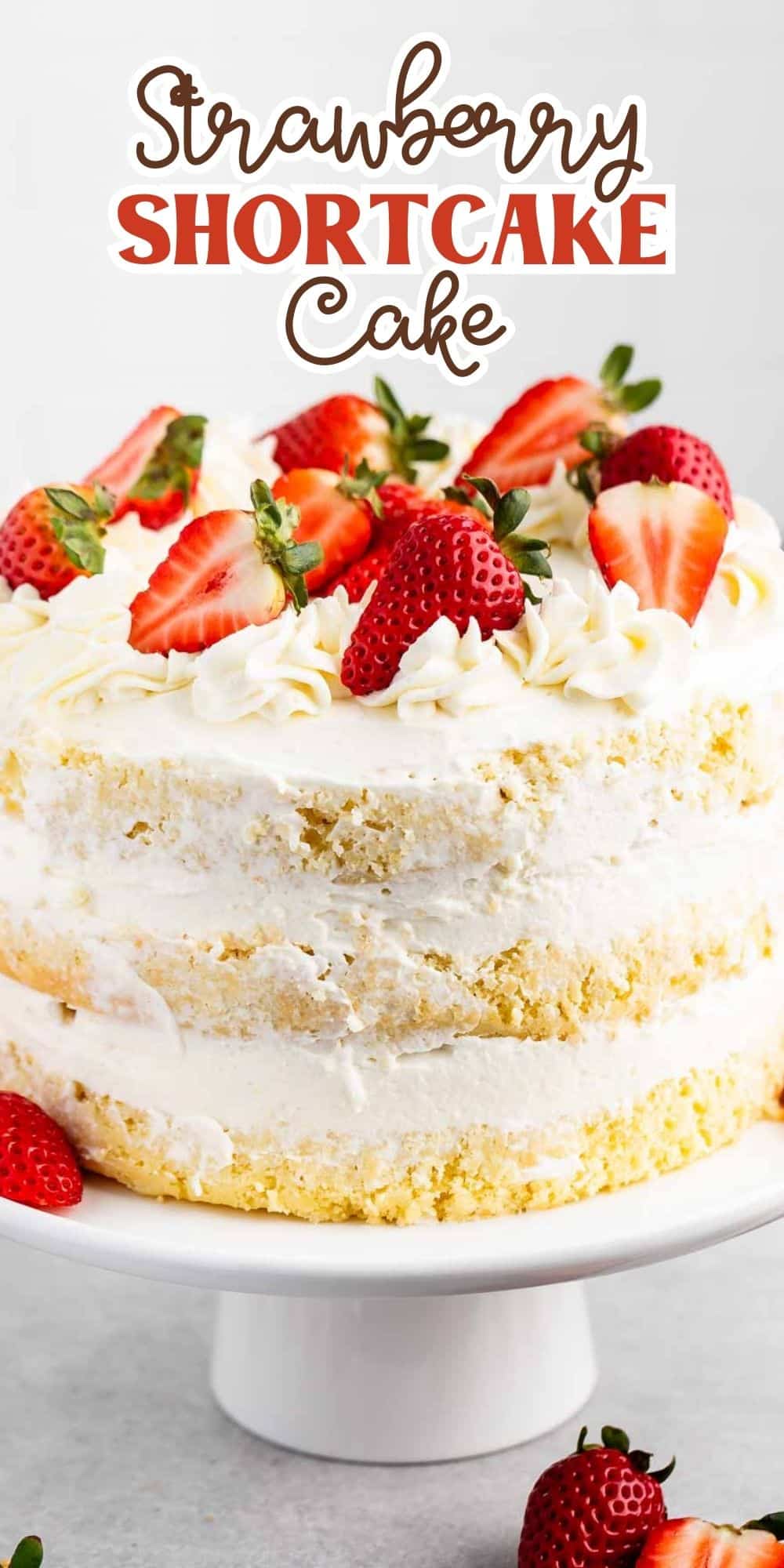 A layered strawberry shortcake with whipped cream and fresh strawberry halves on top. The cake is displayed on a white pedestal, with strawberries scattered nearby. The text above reads Strawberry Shortcake Cake.