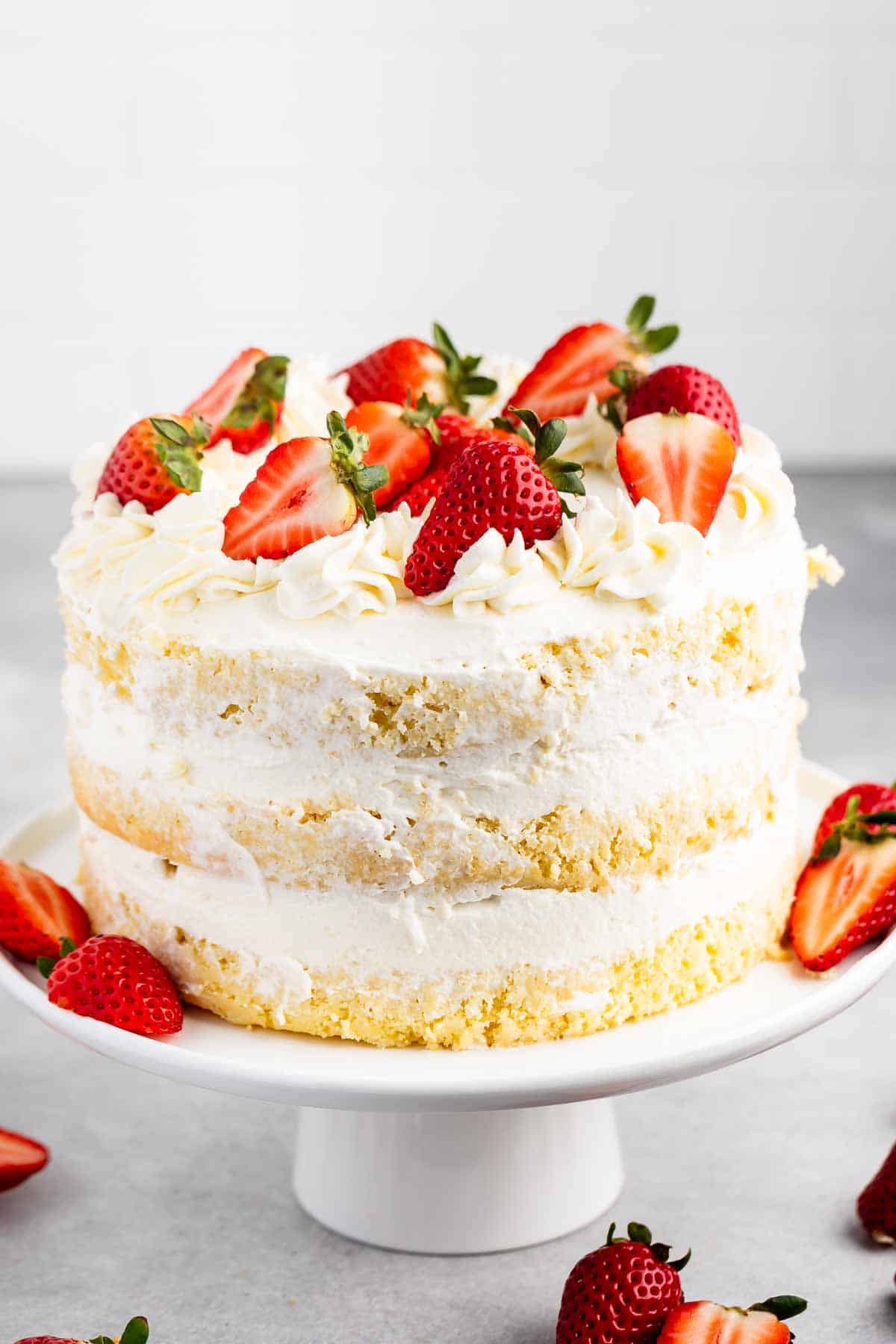 A layered vanilla cake with creamy white frosting, topped with halved strawberries. The cake is displayed on a white pedestal, against a neutral background. Additional strawberries are placed around the base of the stand.