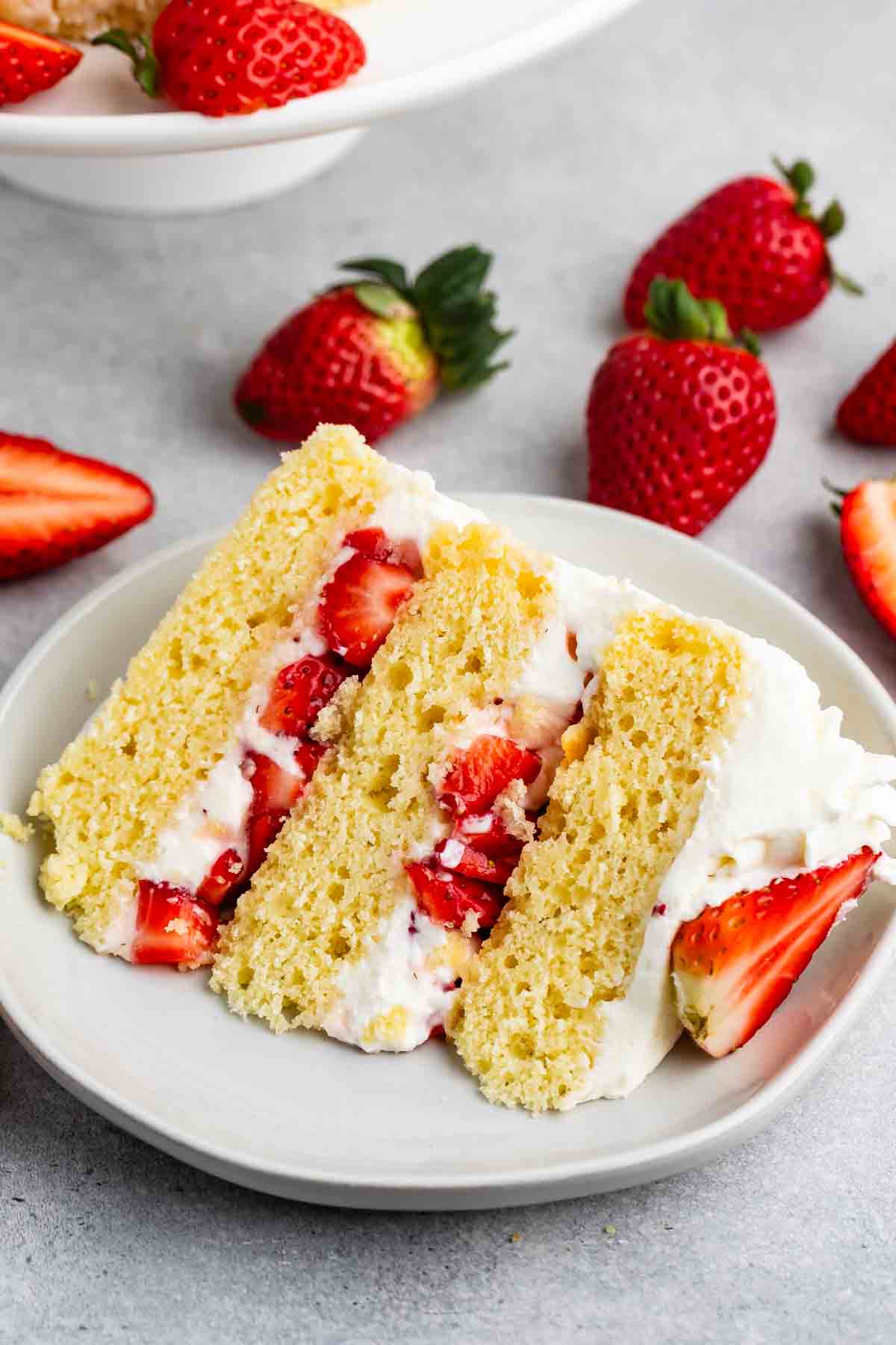 A slice of strawberry shortcake on a white plate. The layered cake is filled with fresh strawberries and cream. Whole strawberries are scattered around the plate on a gray surface. A partially visible whole cake is in the background.