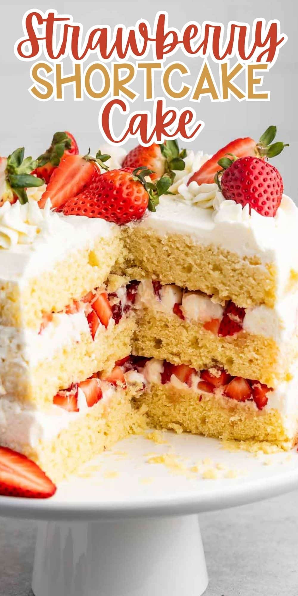 A layer cake sits on a white cake stand. The cake is filled with whipped cream and sliced strawberries between layers of yellow sponge. The top is decorated with strawberries and cream swirls. Text above reads Strawberry Shortcake Cake.