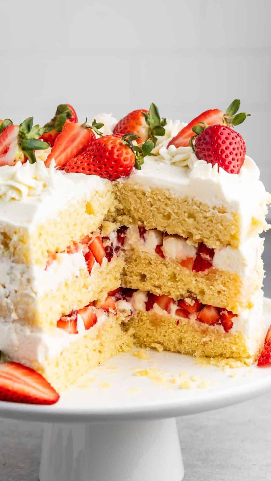 A three-layer cake with white frosting, topped and layered with fresh strawberries. The cake is decorated with whole and sliced strawberries and placed on a round white cake stand.