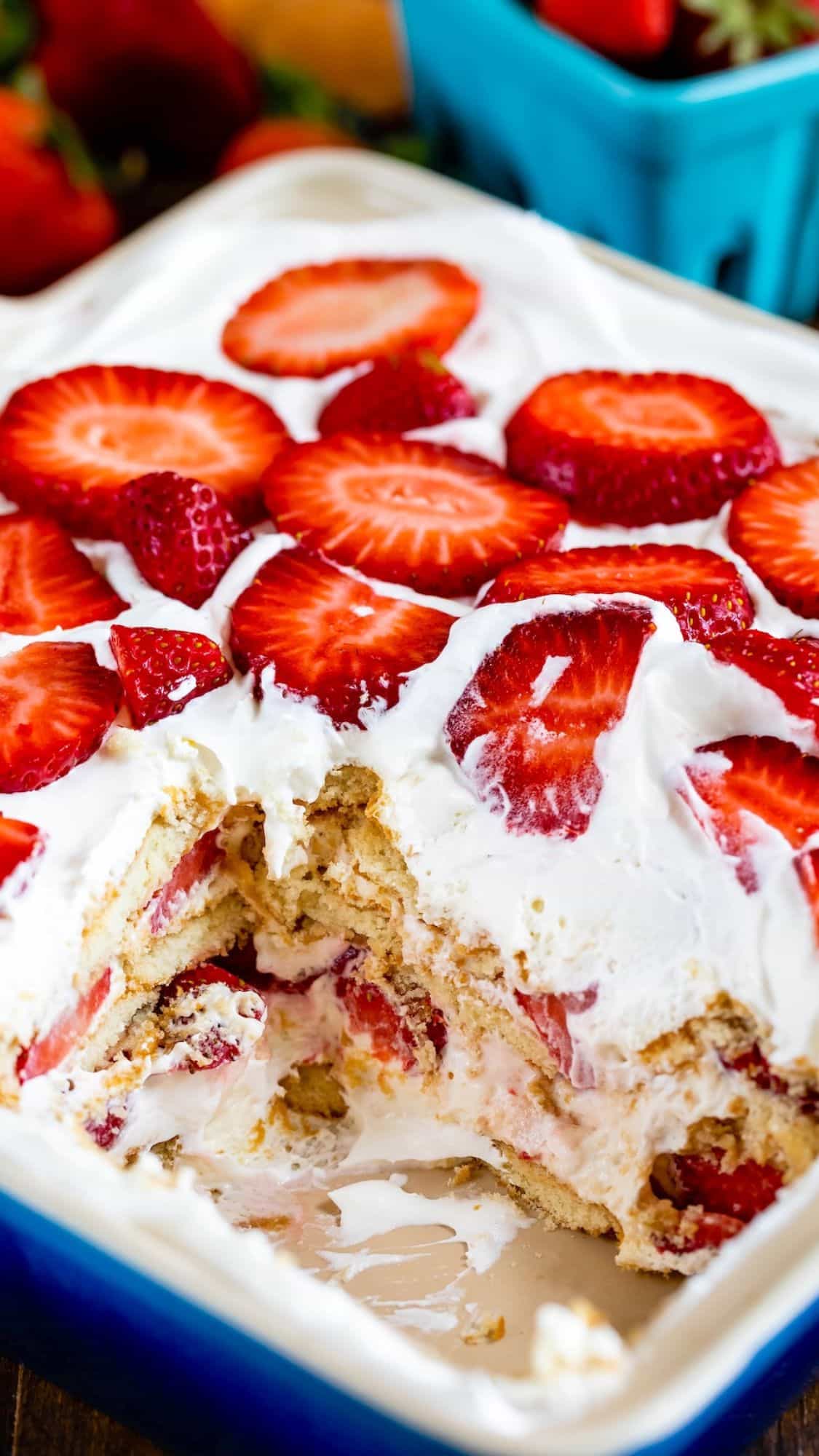 A dessert dish with layers of sliced strawberries and whipped cream, showing a serving removed to reveal layers within. The dish has a blue rim, and there are fresh strawberries in the background.